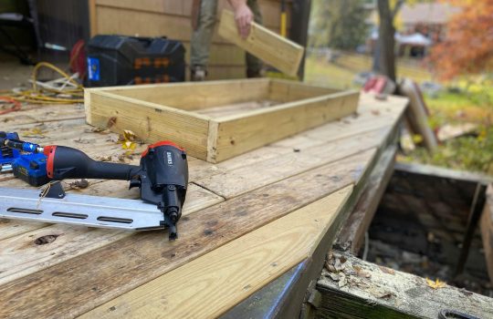 Close-up of carpentry work on a deck with a nail gun and wooden frame under construction.